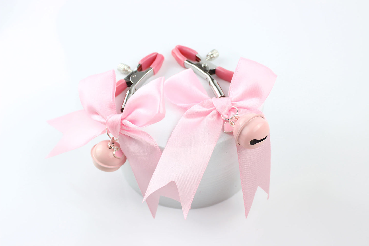 Two metal adjustable screw nipple clamps with pink PVC caps, pink satin bows, pink bells, and pink heart charms laying over a foam circle on a white background