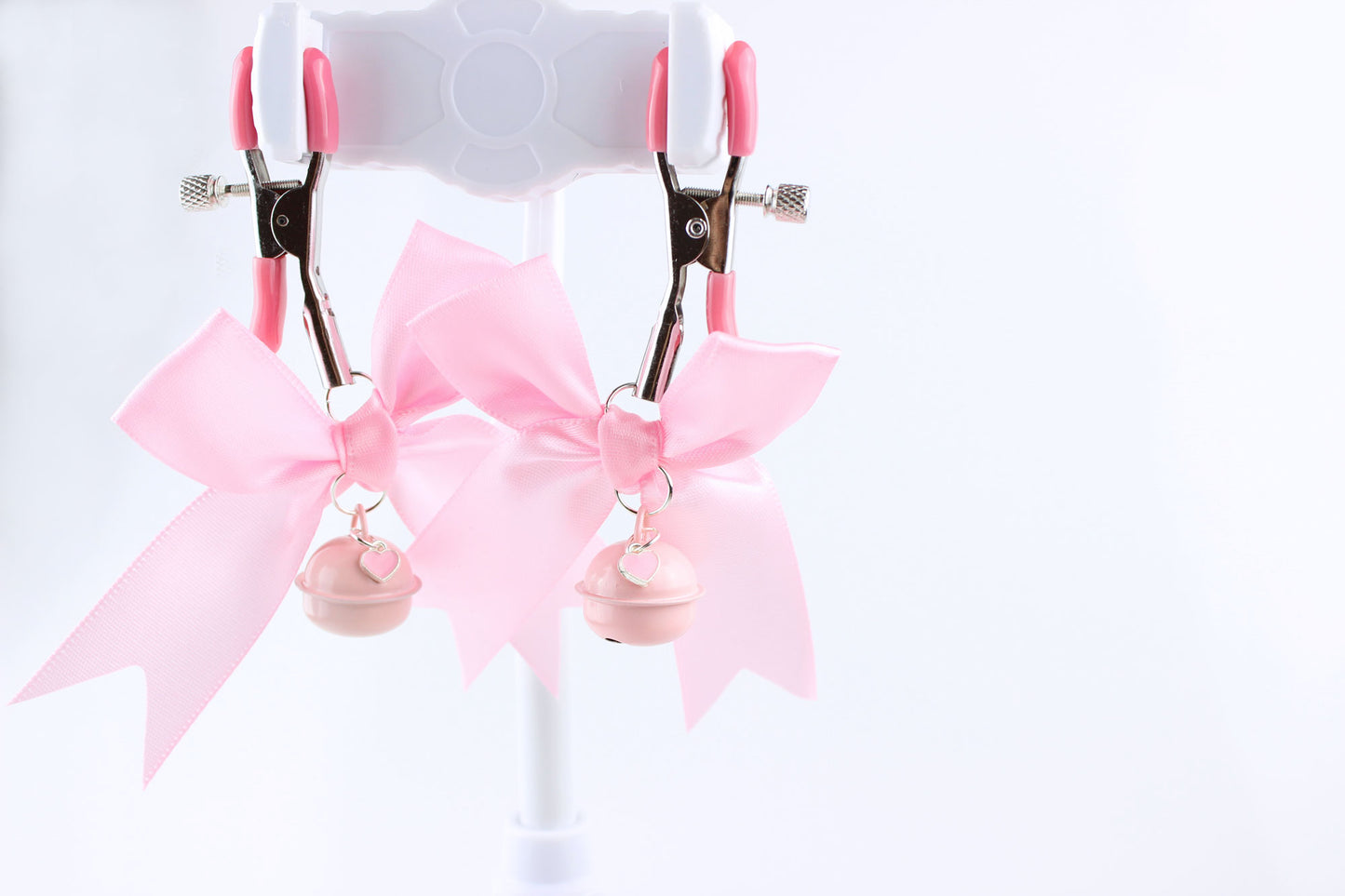 Two metal adjustable screw nipple clamps with pink PVC caps, pink satin bows, pink bells, and pink heart charms hanging in front of a white background