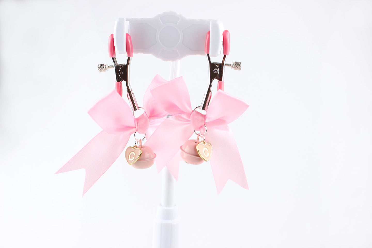 Two adjustable metal nipple clamps with pink PVC caps, satin bows, and heart shaped letter charms in front of a white background