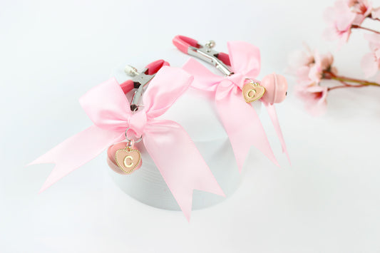 Two adjustable metal nipple clamps with pink PVC caps, satin bows, and heart shaped letter charms on a white background in front of pink flowers