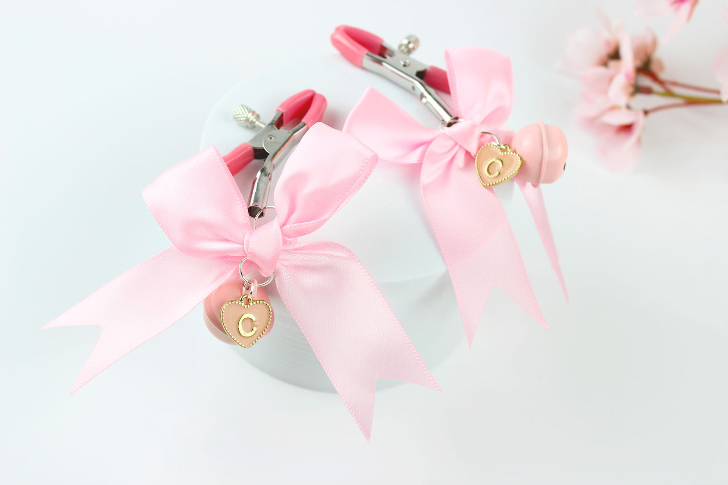 Two adjustable metal nipple clamps with pink PVC caps, satin bows, and heart shaped letter charms on a white background in front of pink flowers