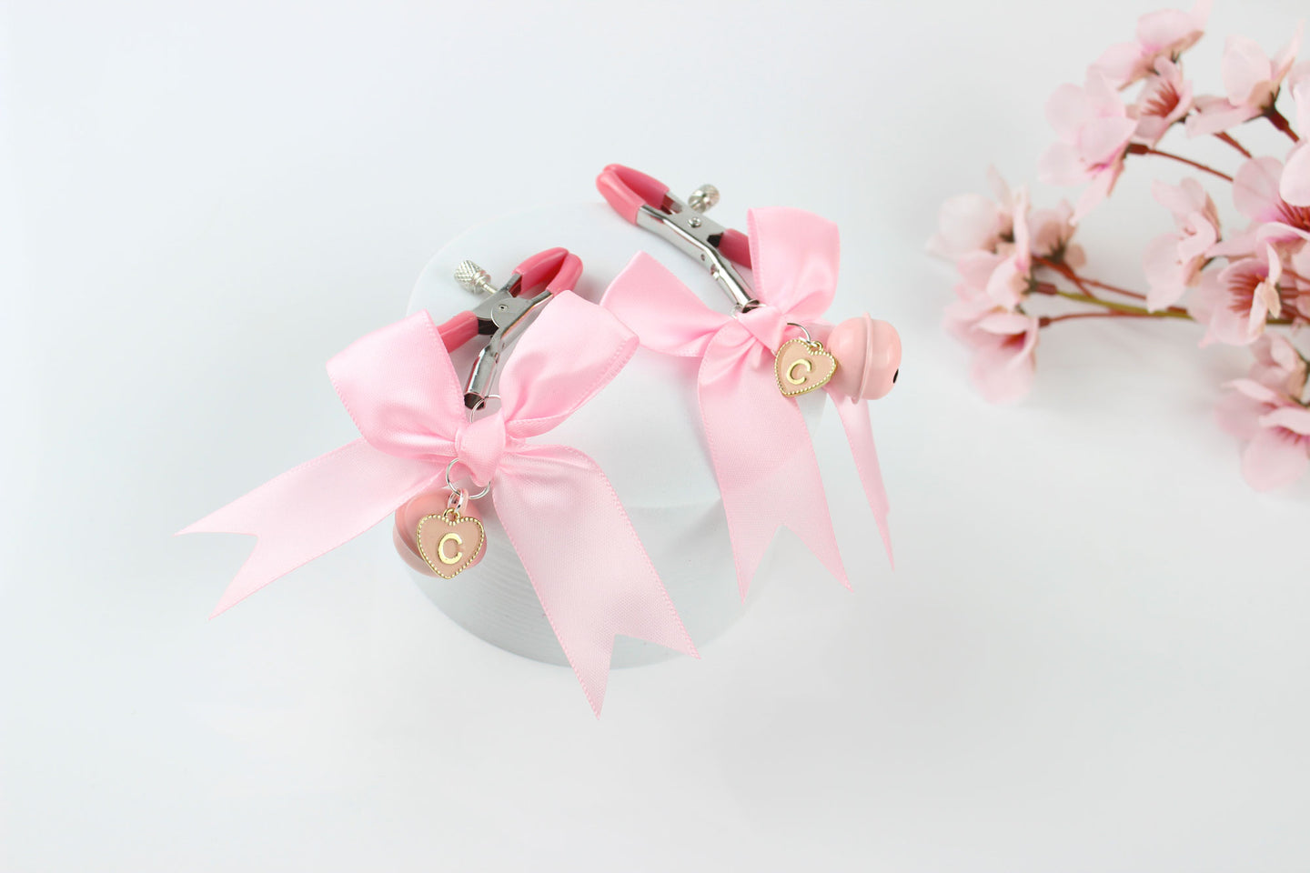 Two adjustable metal nipple clamps with pink PVC caps, satin bows, and heart shaped letter charms on a white background in front of pink flowers