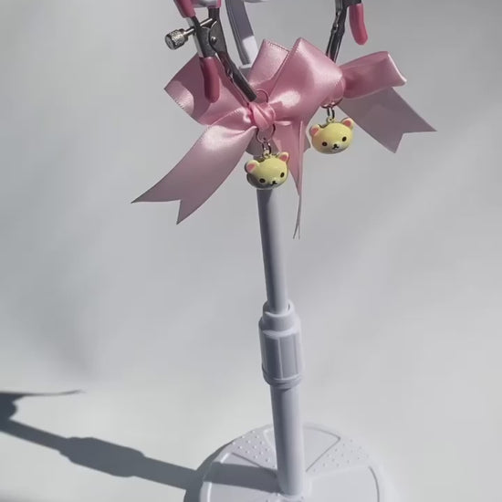 Video of Silver nipple clamps with pink pvc caps and adjustable tension screw and pink bows and white bear heads hanging off the clamps being held up on a white background and rotated around.