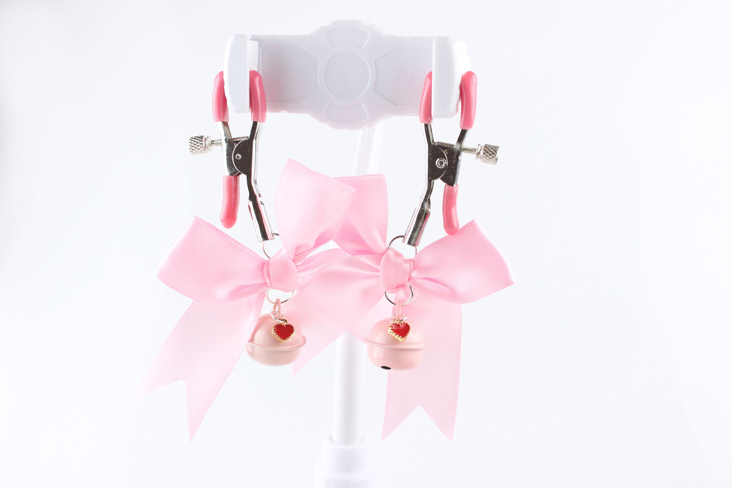 Two metal adjustable screw nipple clamps with pink PVC caps, pink satin bows, pink bells, and red heart charms hanging in front of a white background