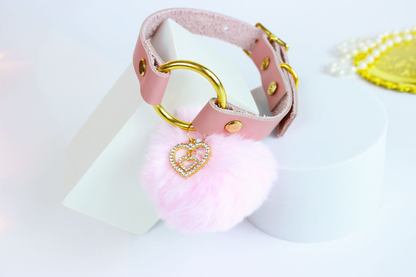 Pink leather collar with gold hardware, pink pom charm, and gold rhinestone heart charm in front of white background