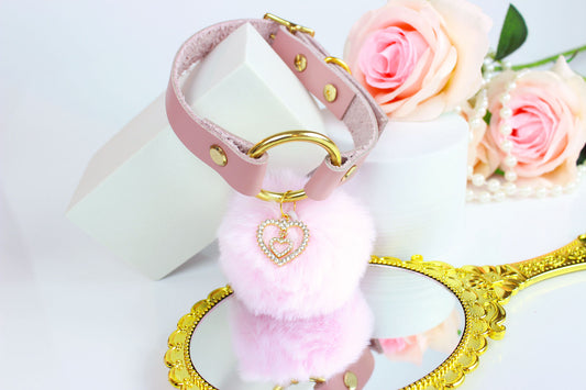 Pink leather collar with gold hardware, pink pom charm, and gold rhinestone heart charm over gold framed hand mirror with roses and pearls in background