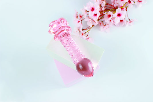 Pink glass dildo with a rose base and a ridged shaft on white square over white background.