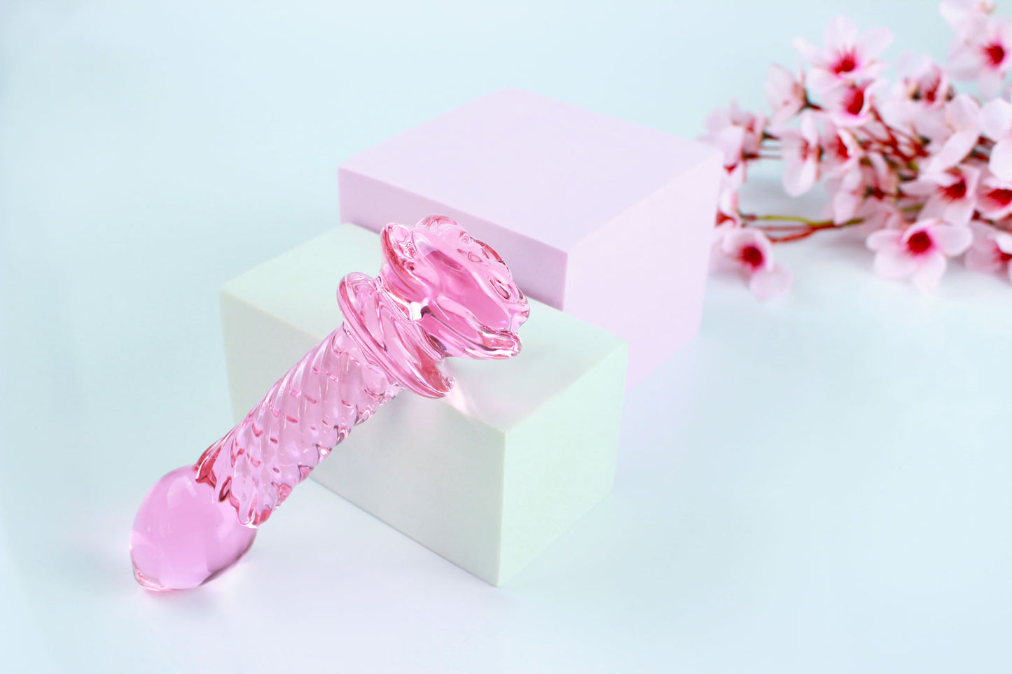 Pink glass dildo with a rose base and a ridged shaft on white square over white background.