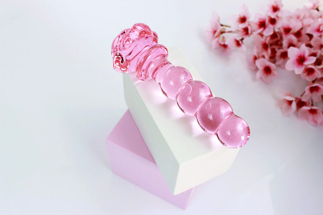 Pink glass dildo with a rose base and a knotted shaft in front of a white backdrop with flowers.