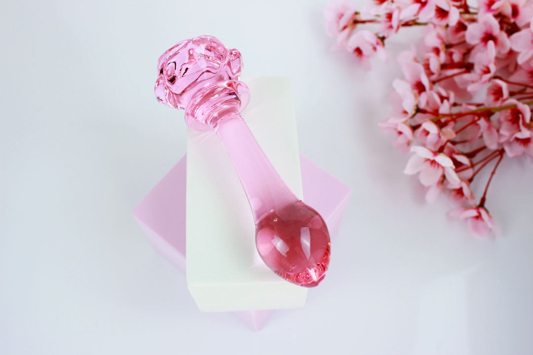 Pink glass butt plug with a rose base elevated on a white square against a white backdrop with flowers.