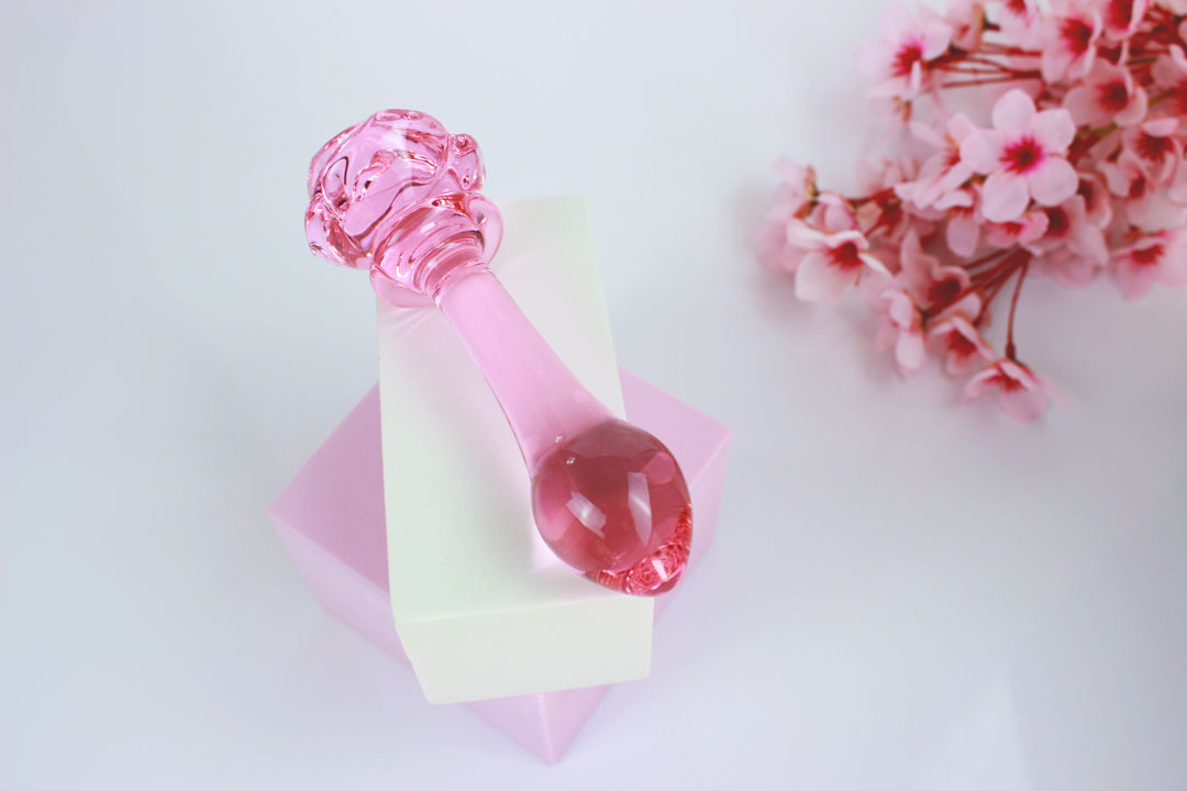 Pink glass butt plug with a rose base elevated on a white square against a white backdrop with flowers.