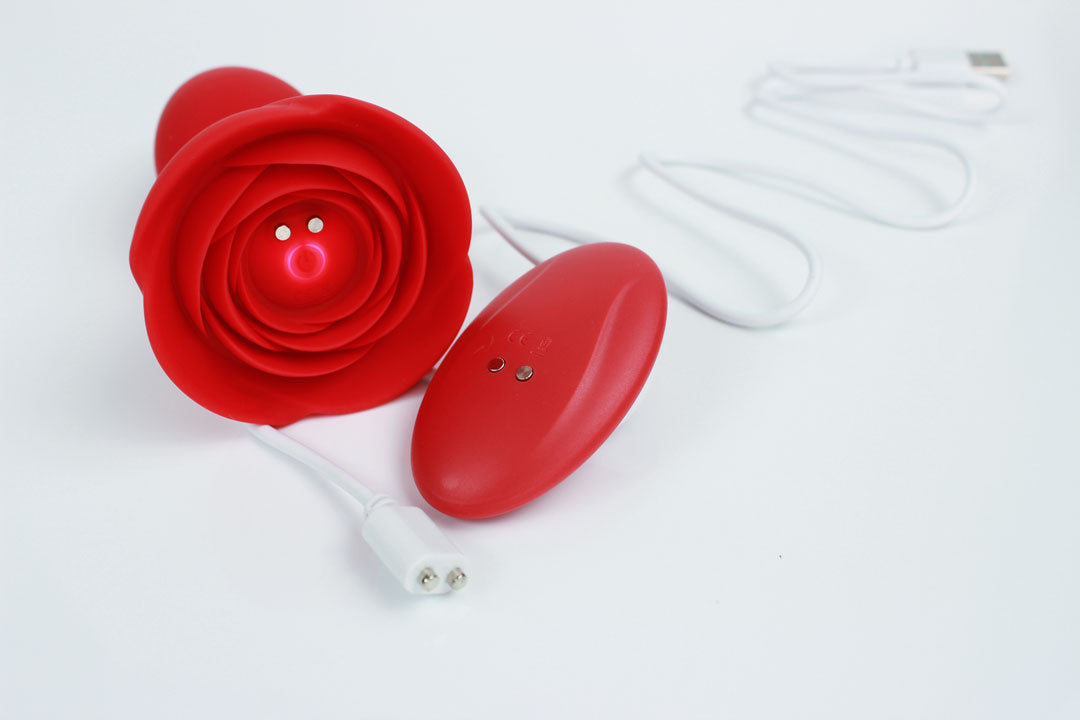 red vibrating butt plug with rose base, remote control, and charging cord on a white background 