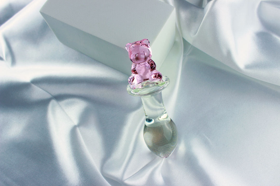 Bear glass butt plug with a bulbed shaft and a pink bear base leaned against a white block on white satin fabric.