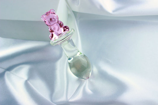 Bear glass butt plug with a bulbed shaft and a pink bear base leaned against a white block on white satin fabric.