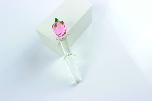 Clear, tapered glass butt plug with pink and green strawberry base on a white square against a white background.