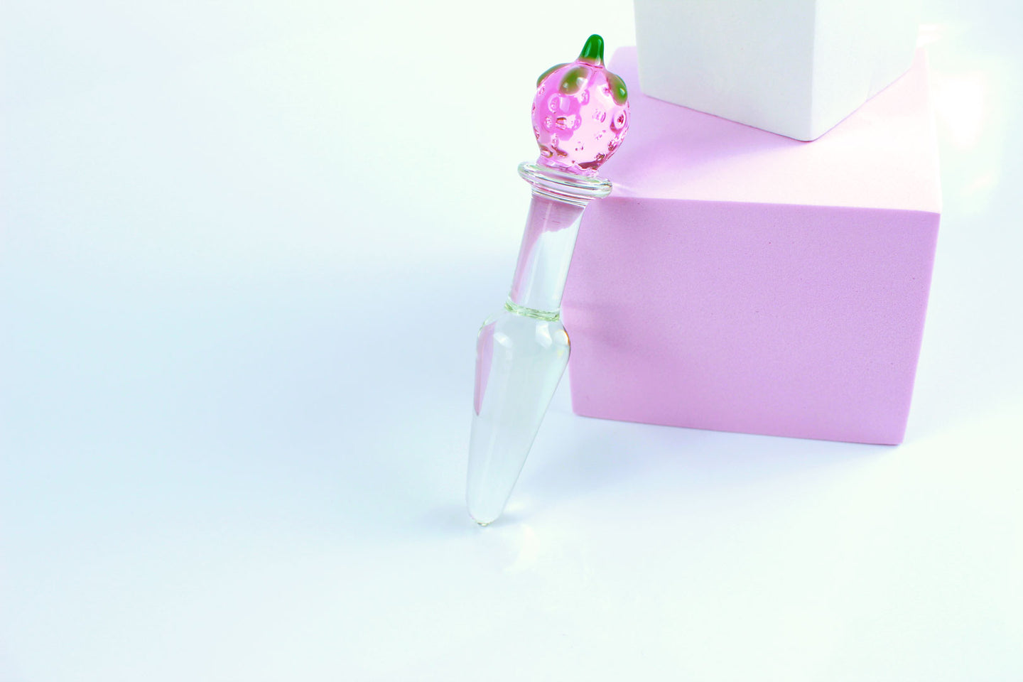 Clear, tapered glass butt plug with pink and green strawberry base laid against a pink block on a white background.