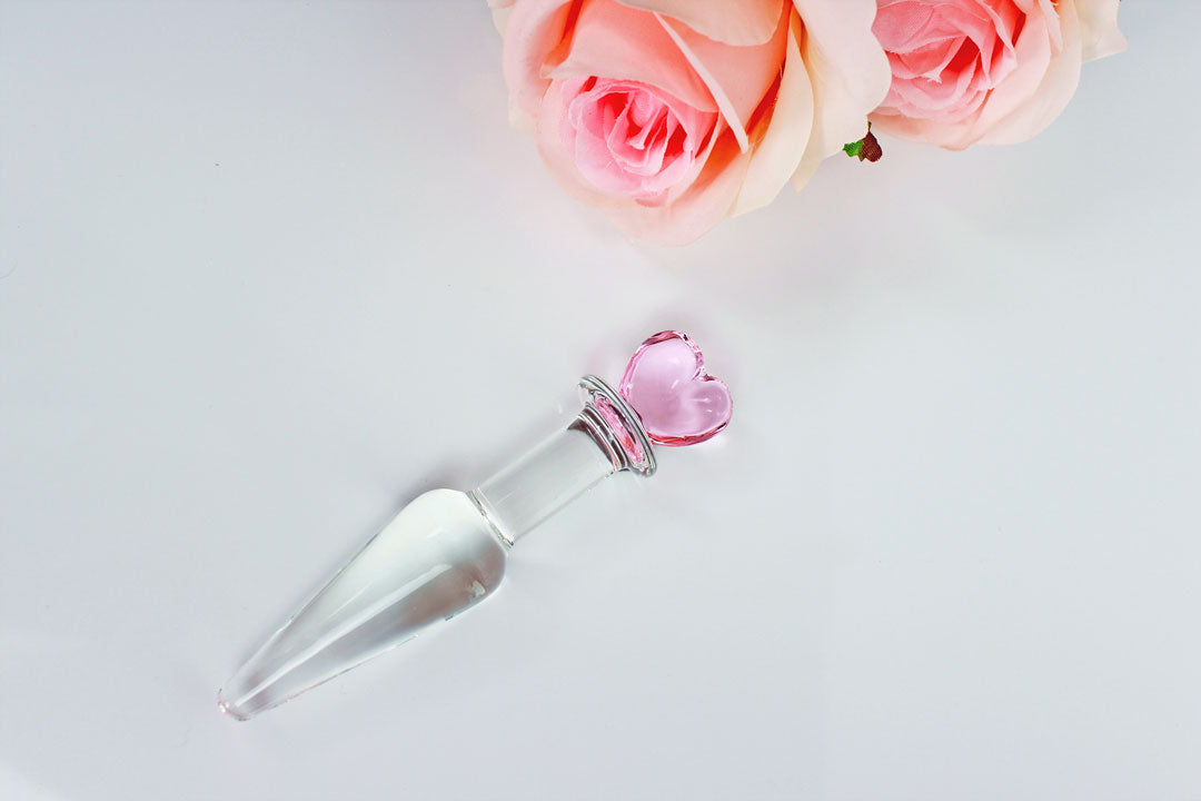 Tapered glass butt plug with pink heart base next to pink roses on a white background.