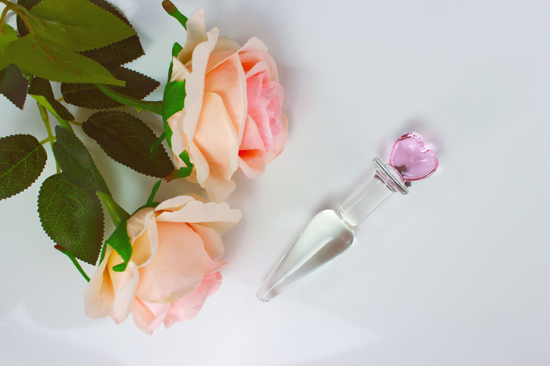 Tapered glass butt plug with pink heart base next to pink roses on a white background.