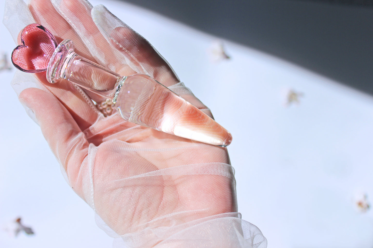 tapered clear butt plug with a pink heart base being held in a gloved hand over a white background with flowers spread