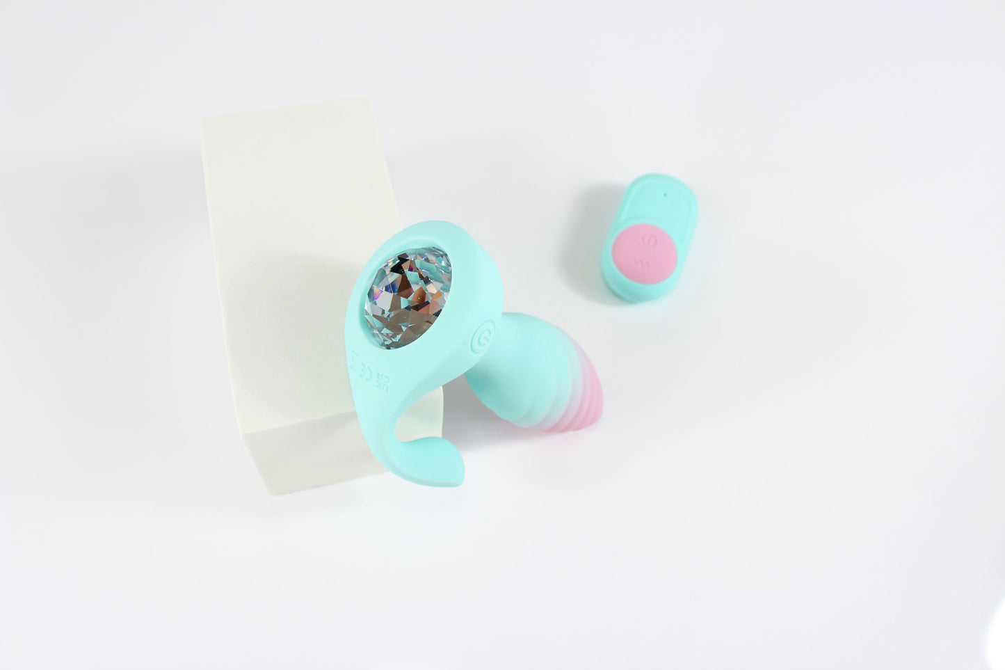 Aerial shot of pastel blue and pink vibrating gem butt plug propped against white block with remote on white background