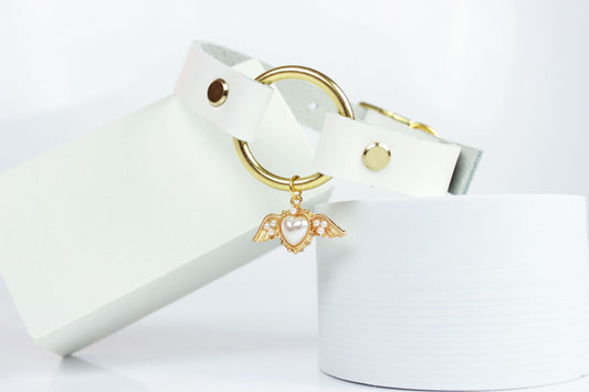 White leather sub collar with gold hardware and pearl winged heart charm in front of white background