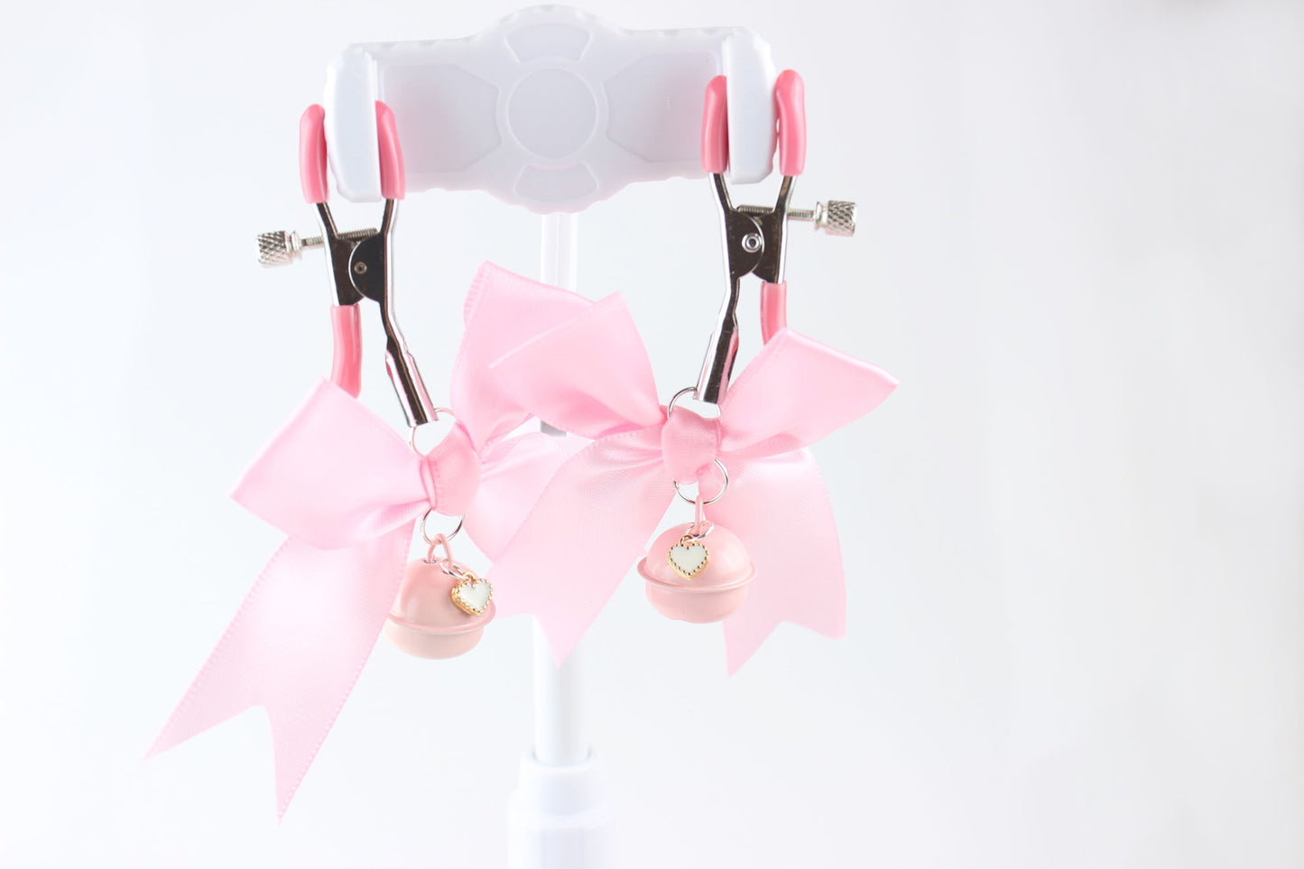 Two metal adjustable screw nipple clamps with pink PVC caps, pink satin bows, pink bells, and white heart charms hanging in front of a white background