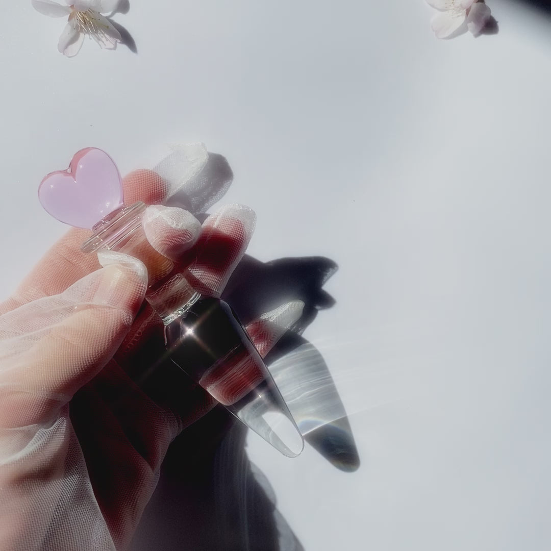 tapered clear glass butt plug with a pink heart base being held in a gloved hand over a white background