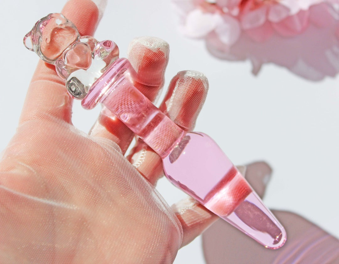 Bear glass butt plug with a pink tapered shaft and clear bear base being held against a white background with flowers.