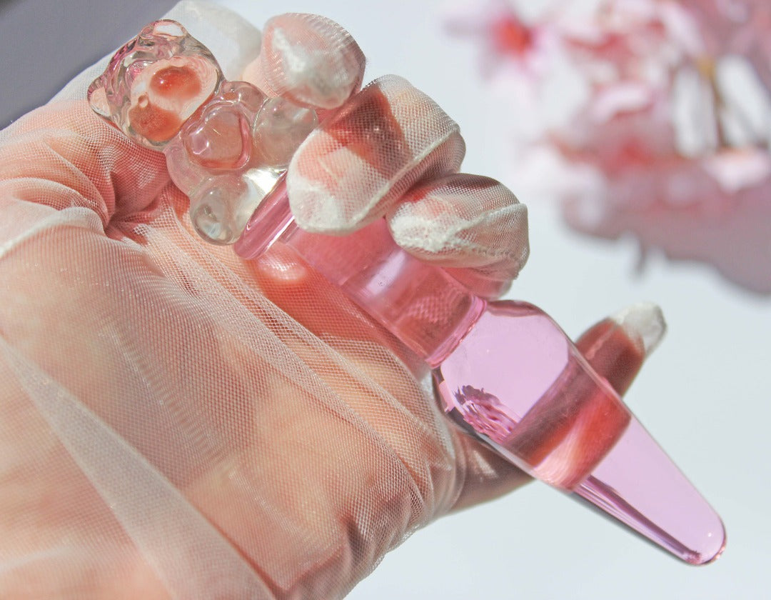 Bear glass butt plug with a pink tapered shaft and clear bear base being held against a white background with flowers.