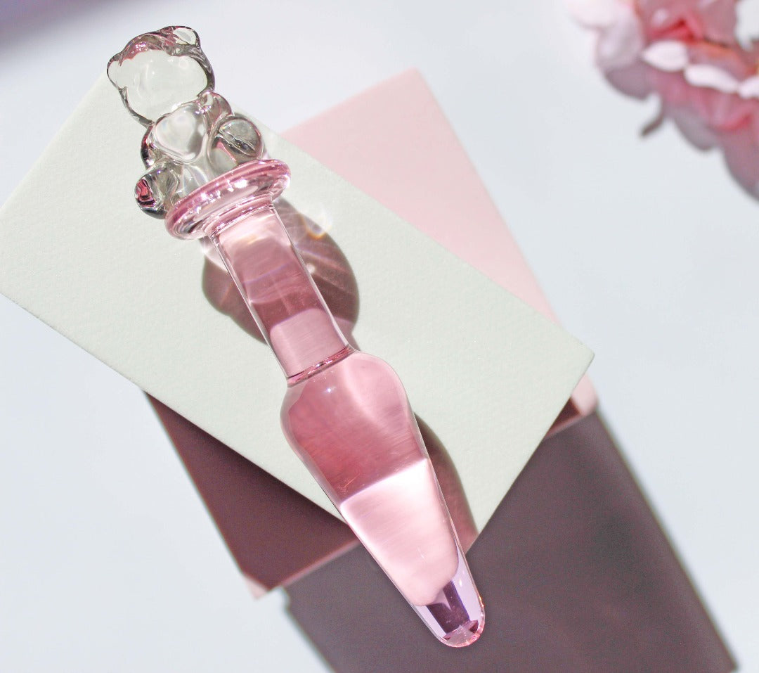 Bear glass butt plug with a pink tapered shaft and clear bear base on white and pink squares on a white background with flowers.