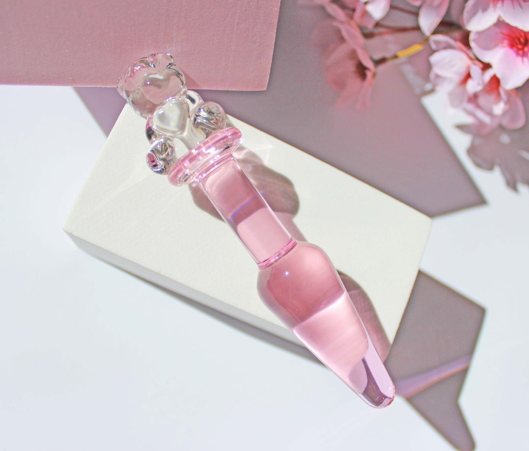 Bear glass butt plug with a pink tapered shaft and clear bear base on white and pink squares on a white background with flowers.