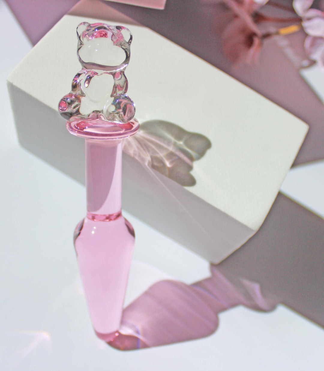 Bear glass butt plug upright with a pink tapered shaft and clear bear base against white block on a white background with flowers.