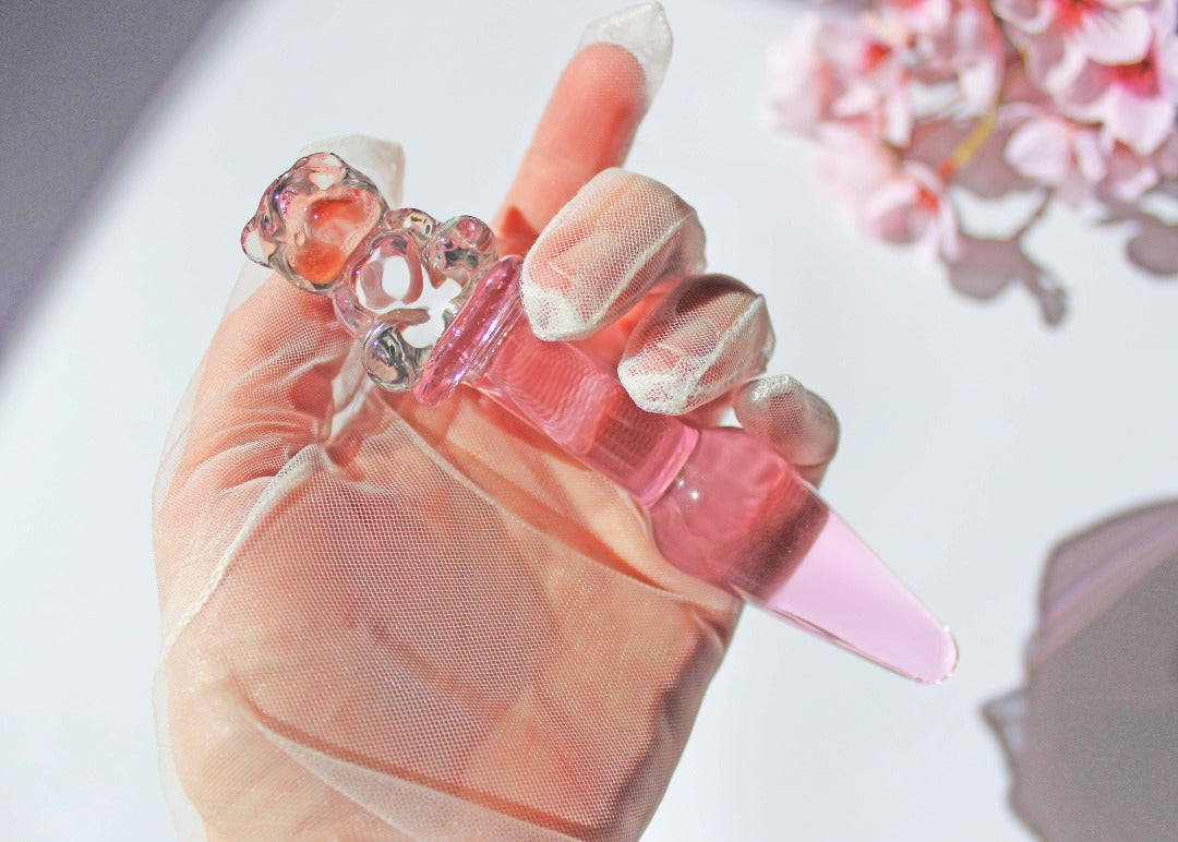 Bear glass butt plug with a pink tapered shaft and clear bear base being held against a white background with flowers.