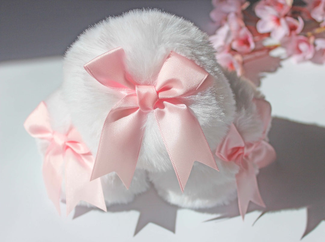 Set of three cute white bunny tail glass butt plugs with pink bows on the pom against a white background with pink flowers.