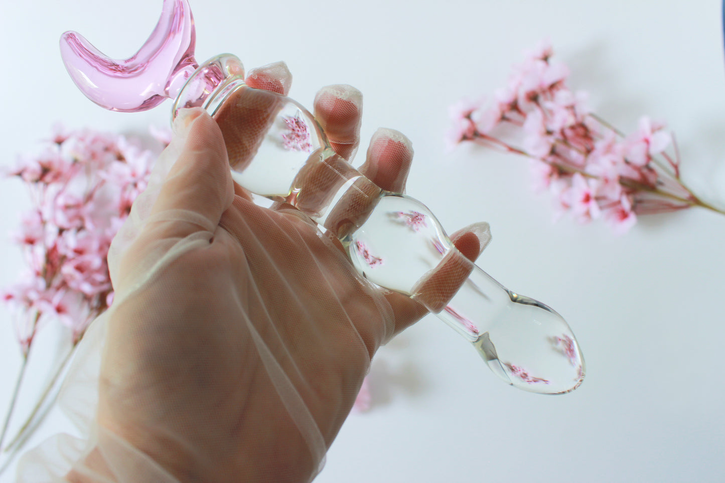 Glass dildo with clear knotted shaft and a pink moon base being held by a gloved hand against a white background with flowers.