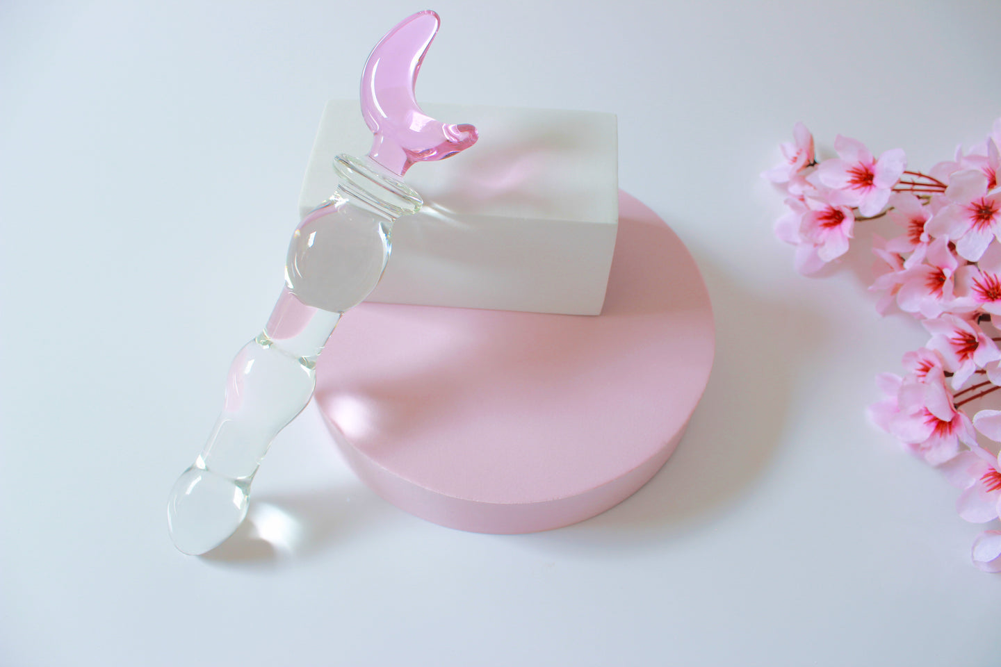 Glass dildo with clear knotted shaft and a pink moon base leaned against a white block on a white background with flowers.