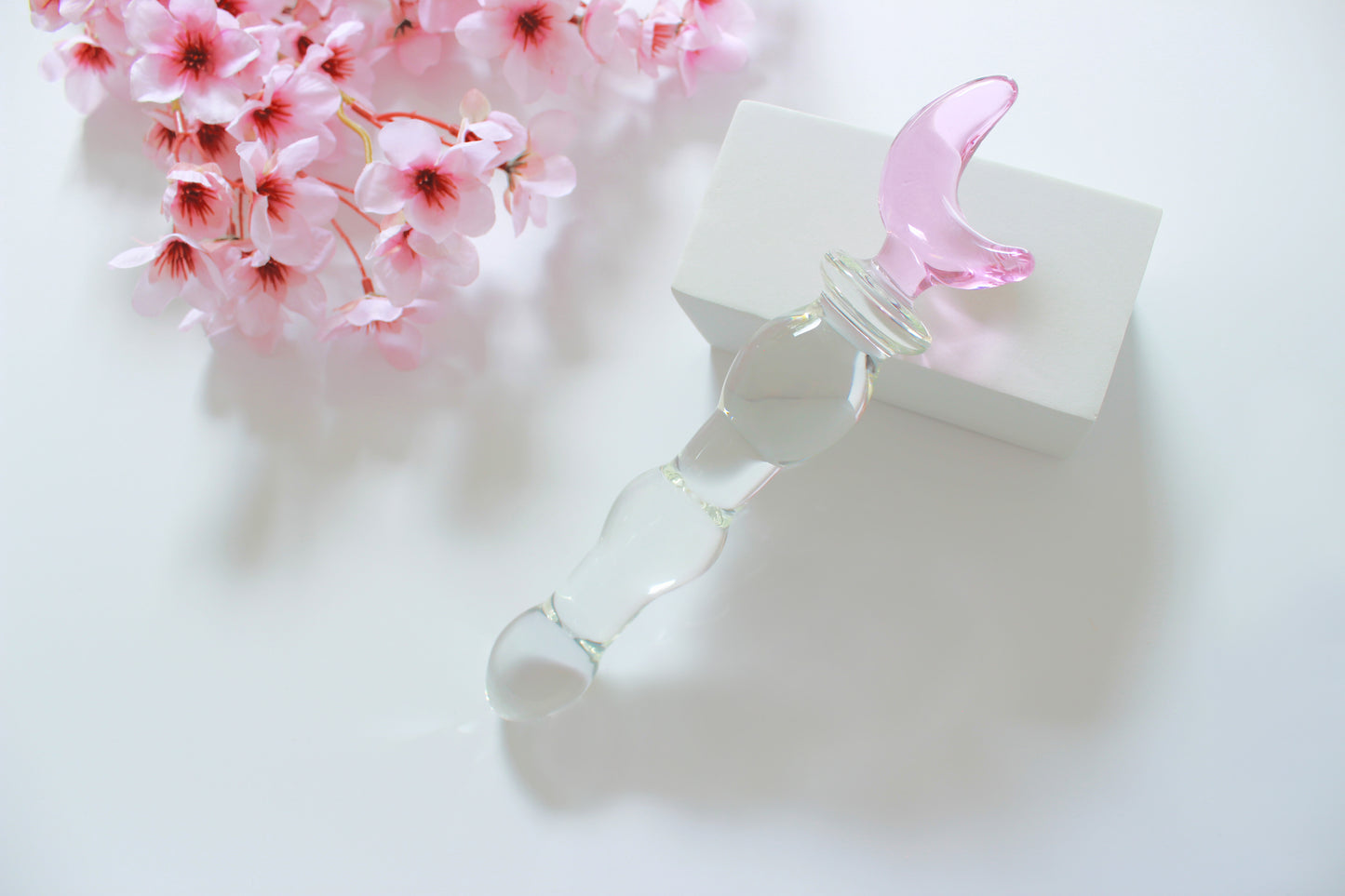 Glass dildo with clear knotted shaft and a pink moon base leaned against a white block on a white background with flowers.