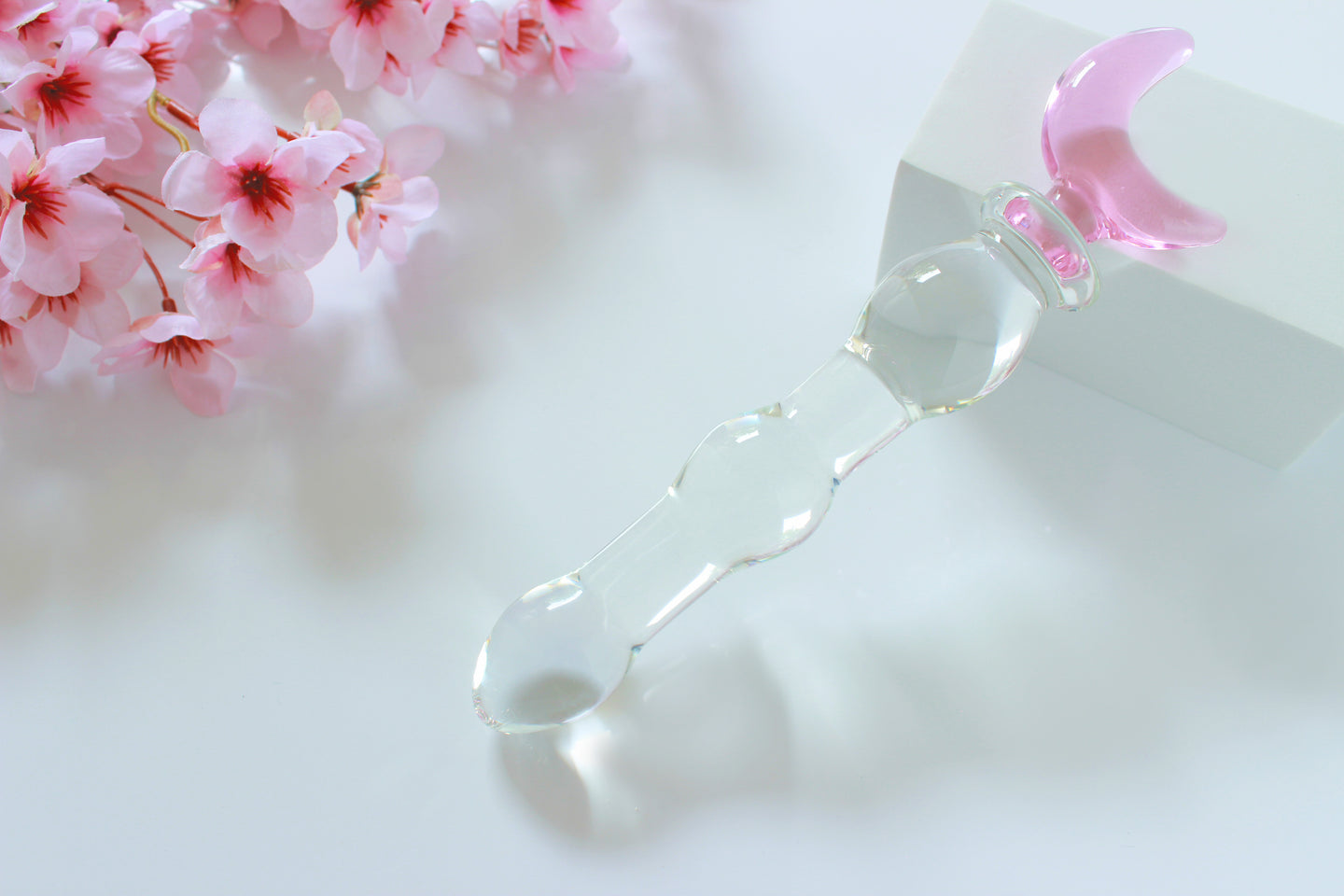 Glass dildo with clear knotted shaft and a pink moon base leaned against a white block on a white background with flowers.