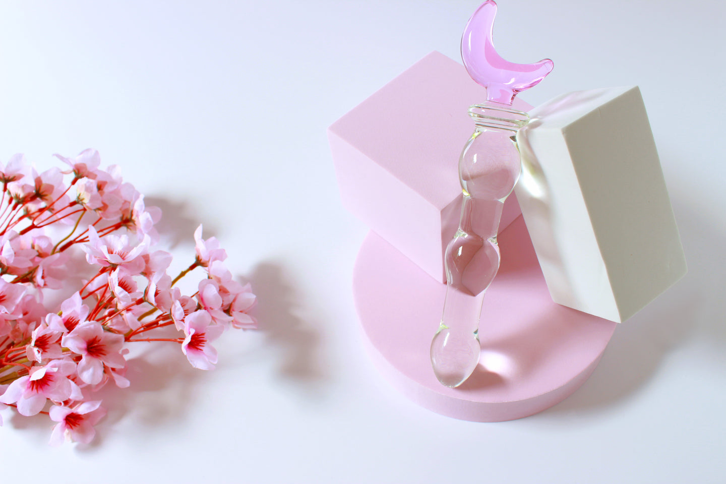 Glass dildo with clear knotted shaft and a pink moon base leaned against a white block on a white background with flowers.