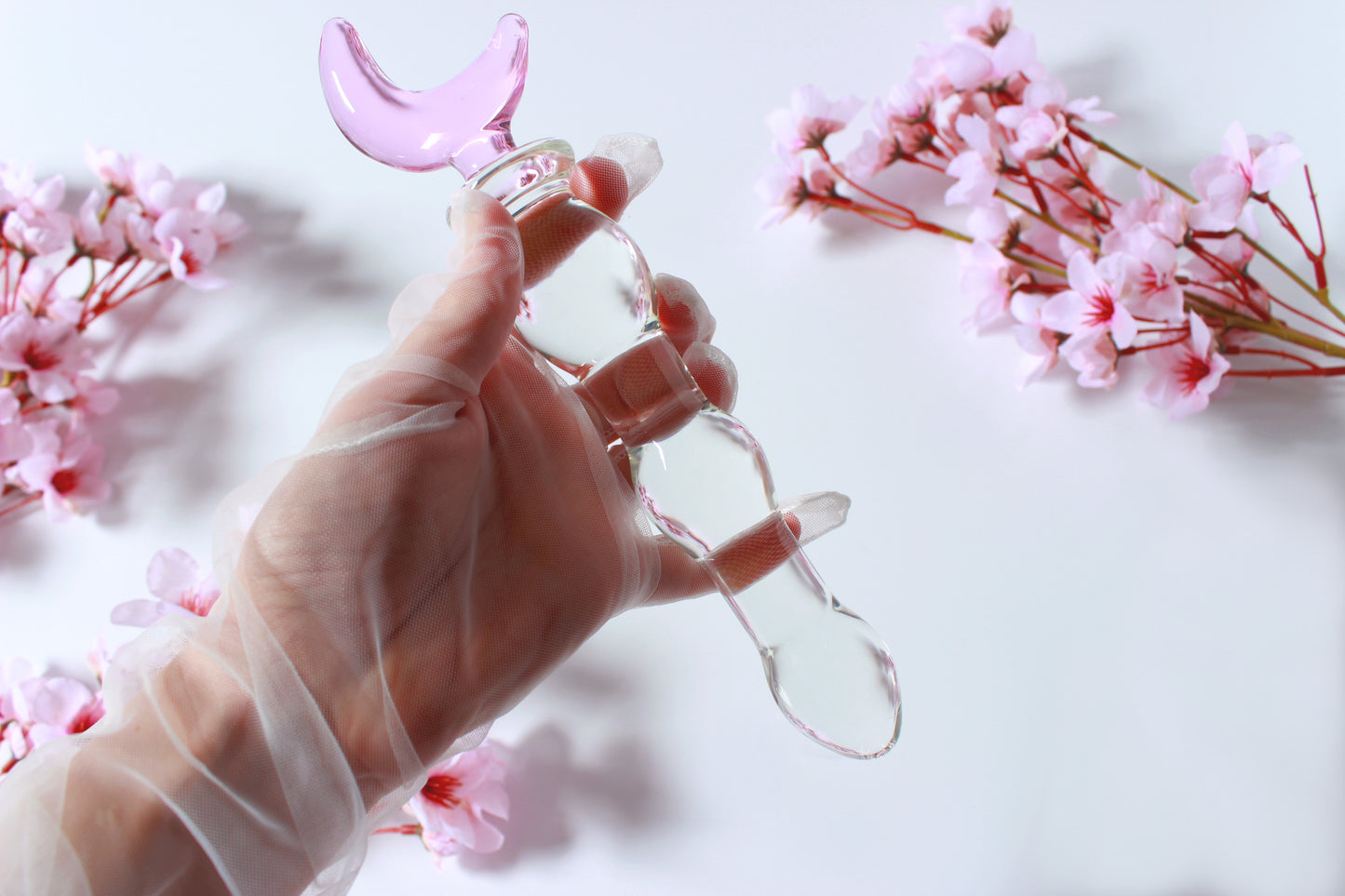 Glass dildo with clear knotted shaft and a pink moon base being held by a gloved hand against a white background with flowers.