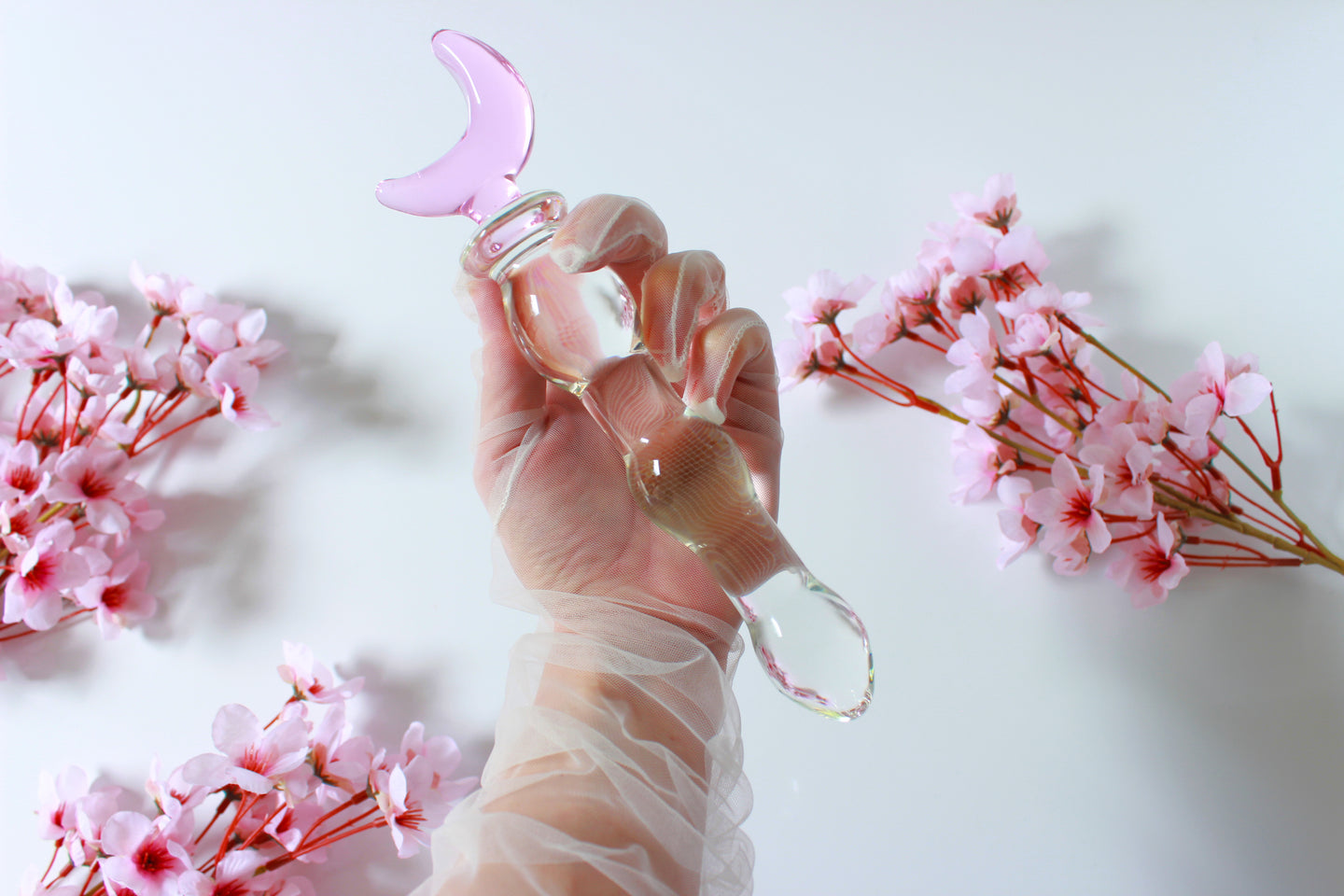 Glass dildo with clear knotted shaft and a pink moon base being held by a gloved hand against a white background with flowers.