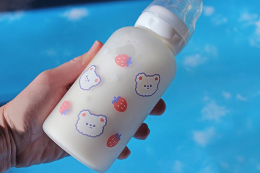 ABDL Adult baby bottle with a white lid and multiple cute white bear heads and strawberries printed on the bottle being held in front of a sky background.