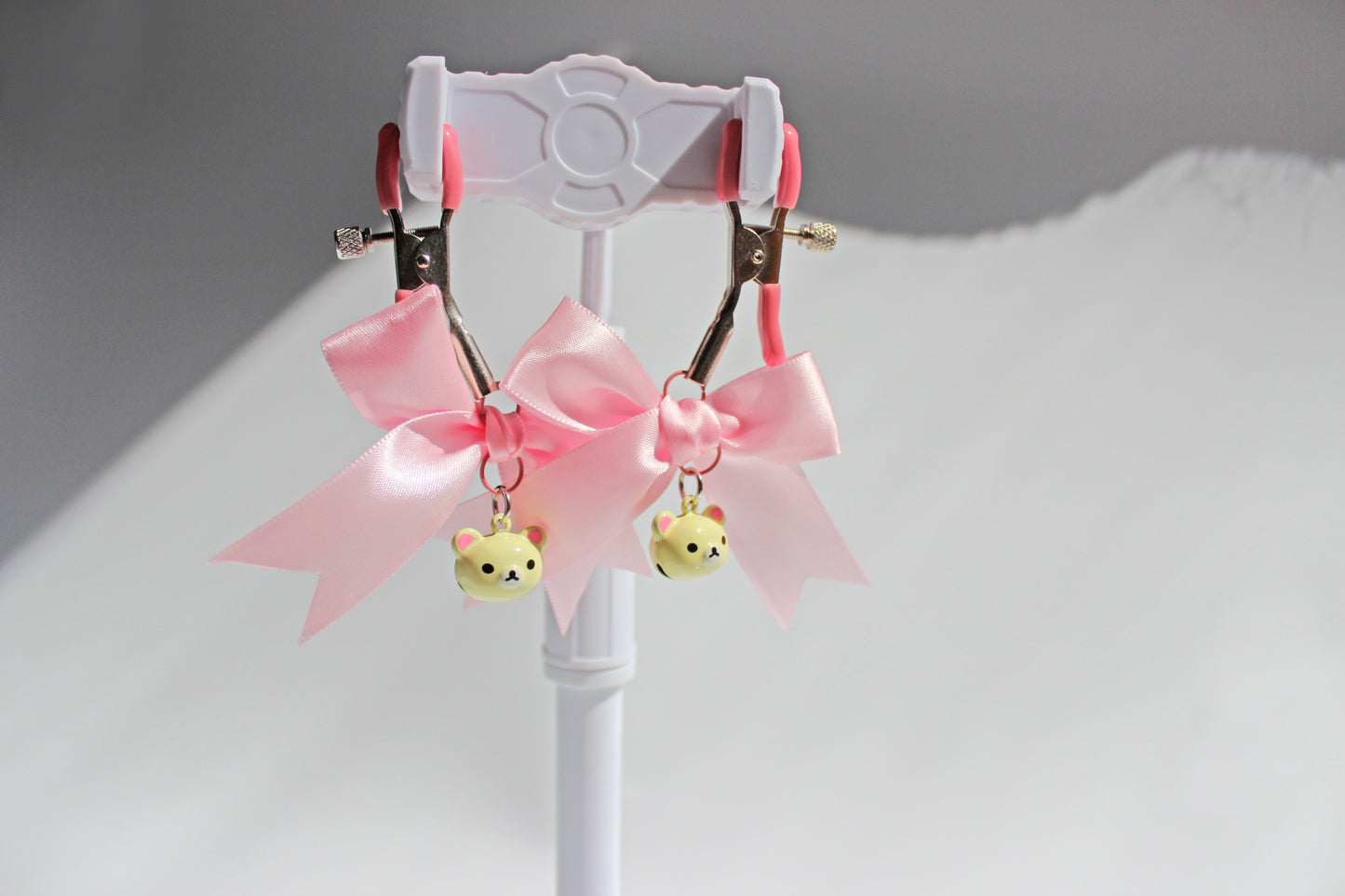 Silver nipple clamps with pink pvc caps and adjustable tension screw and pink bows and white bear heads hanging off the clamps being held up on a white background.