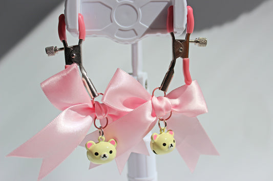 Silver nipple clamps with pink pvc caps and adjustable tension screw and pink bows and white bear heads hanging off the clamps being held up on a white background.