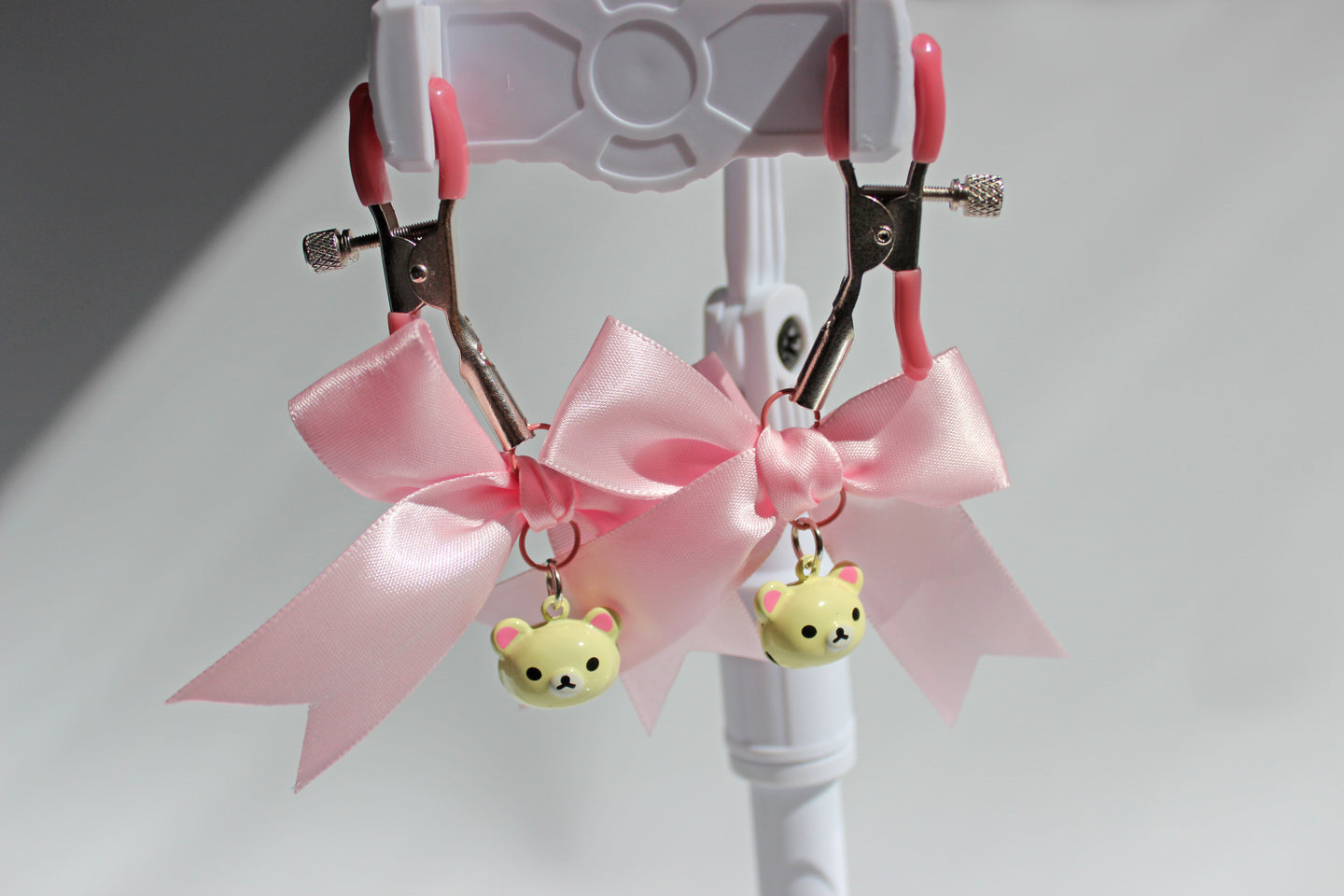 Silver nipple clamps with pink pvc caps and adjustable tension screw and pink bows and white bear heads hanging off the clamps being held up on a white background.