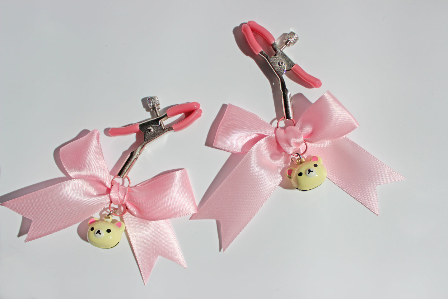 Silver nipple clamps with pink pvc caps and adjustable tension screw and pink bows and white bear heads hanging off the clamps on a white background.