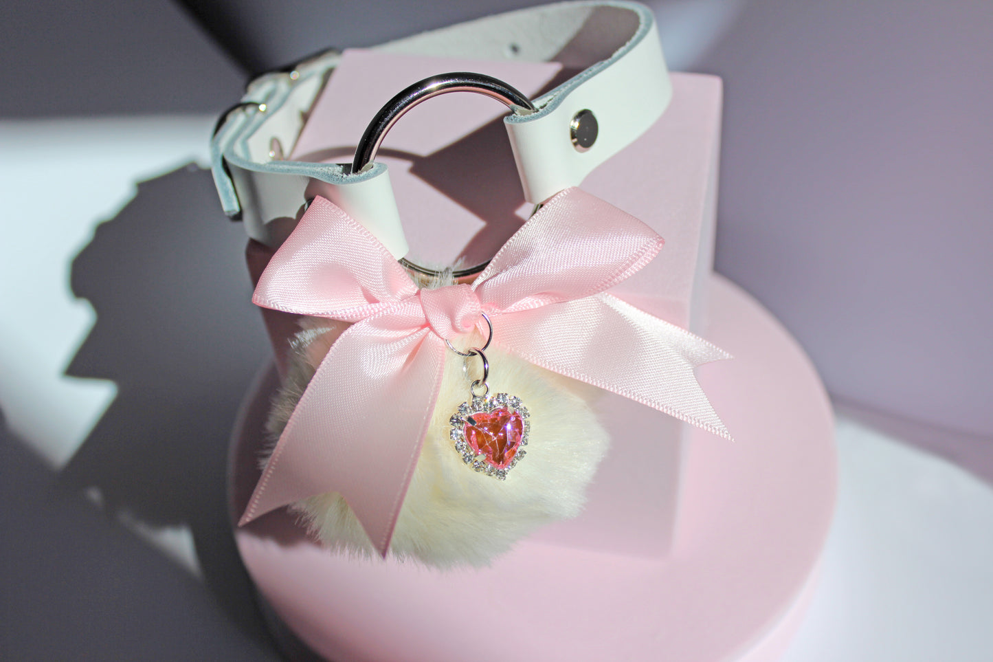 Kawaii white pom collar with a pink bow, heart, and white pom on a pink stand with a white background.