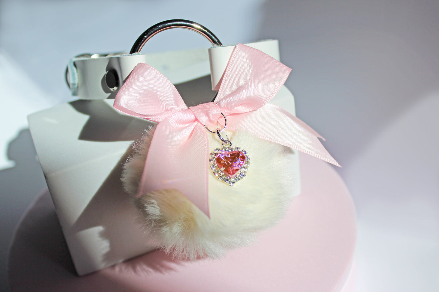 Kawaii white pom collar with a pink bow, heart, and white pom on a pink stand with a white background.