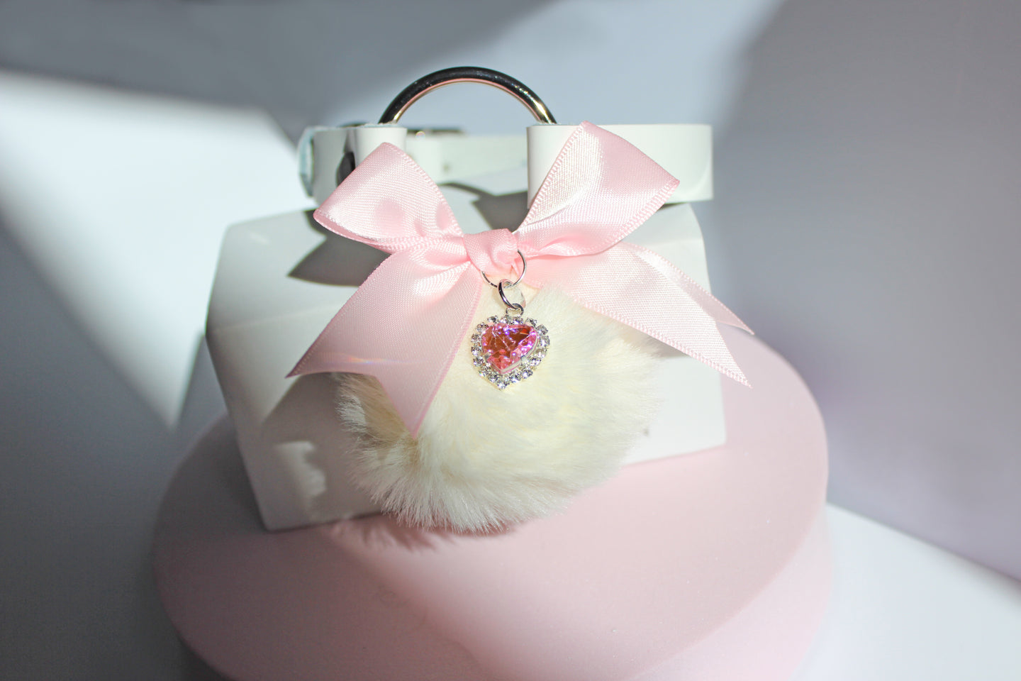 Kawaii white pom collar with a pink bow, heart, and white pom on a pink stand with a white background.