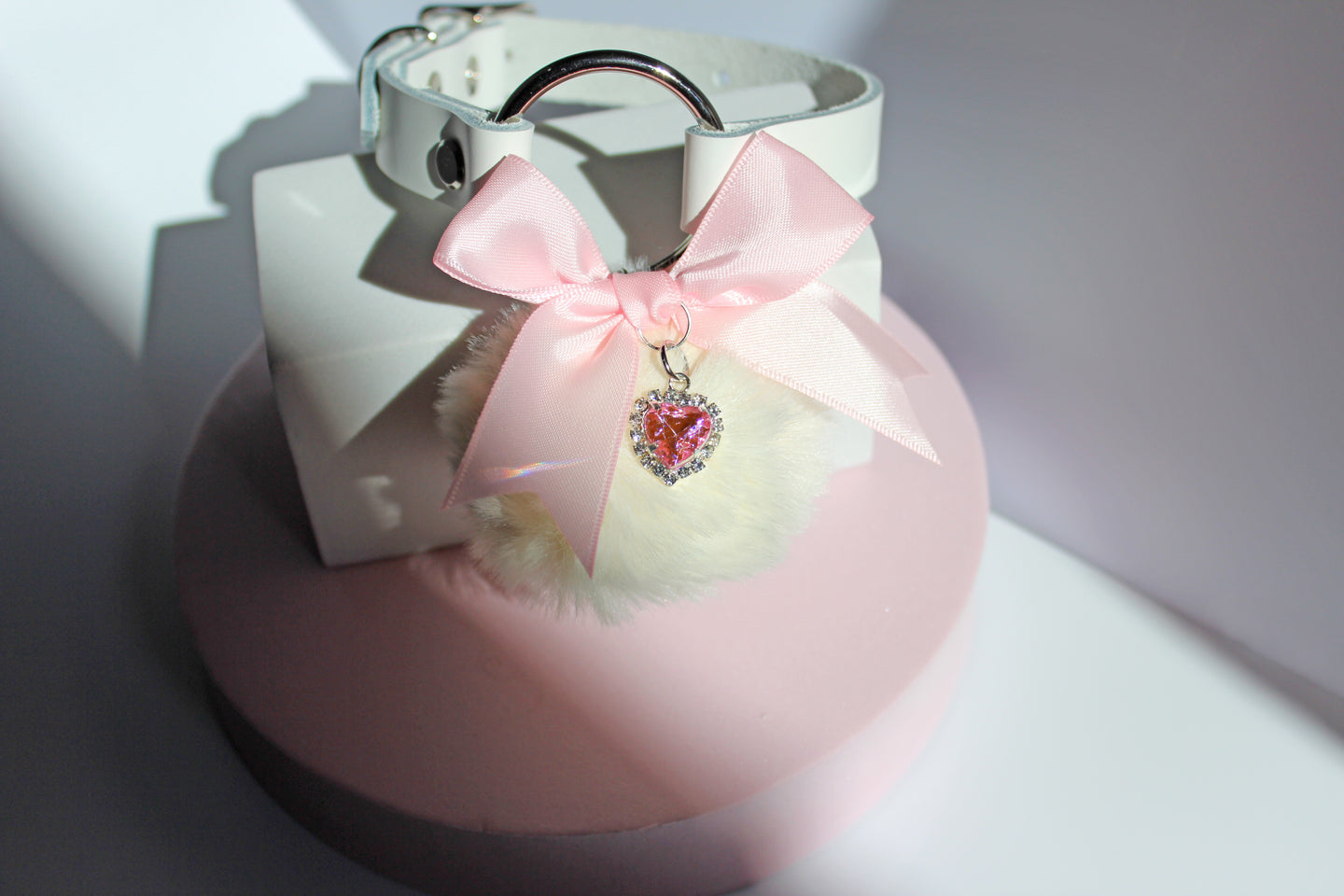 Kawaii white pom collar with a pink bow, heart, and white pom on a pink stand with a white background.